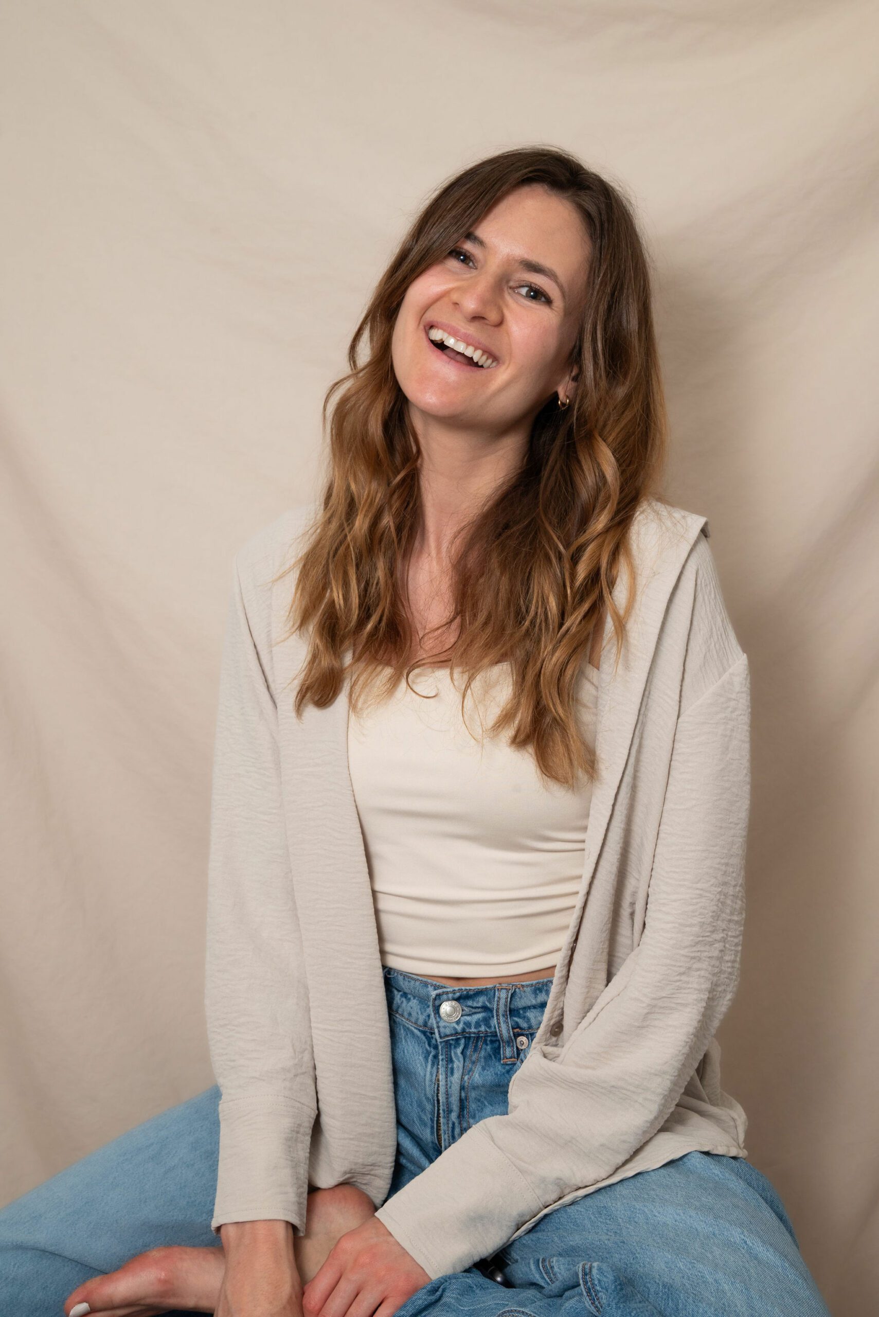 Portrait de l'artiste aquarelle Roxanne Bédard, assise sur un tabouret, souriant, nu pied, en jeans et chemise, devant fond beige.